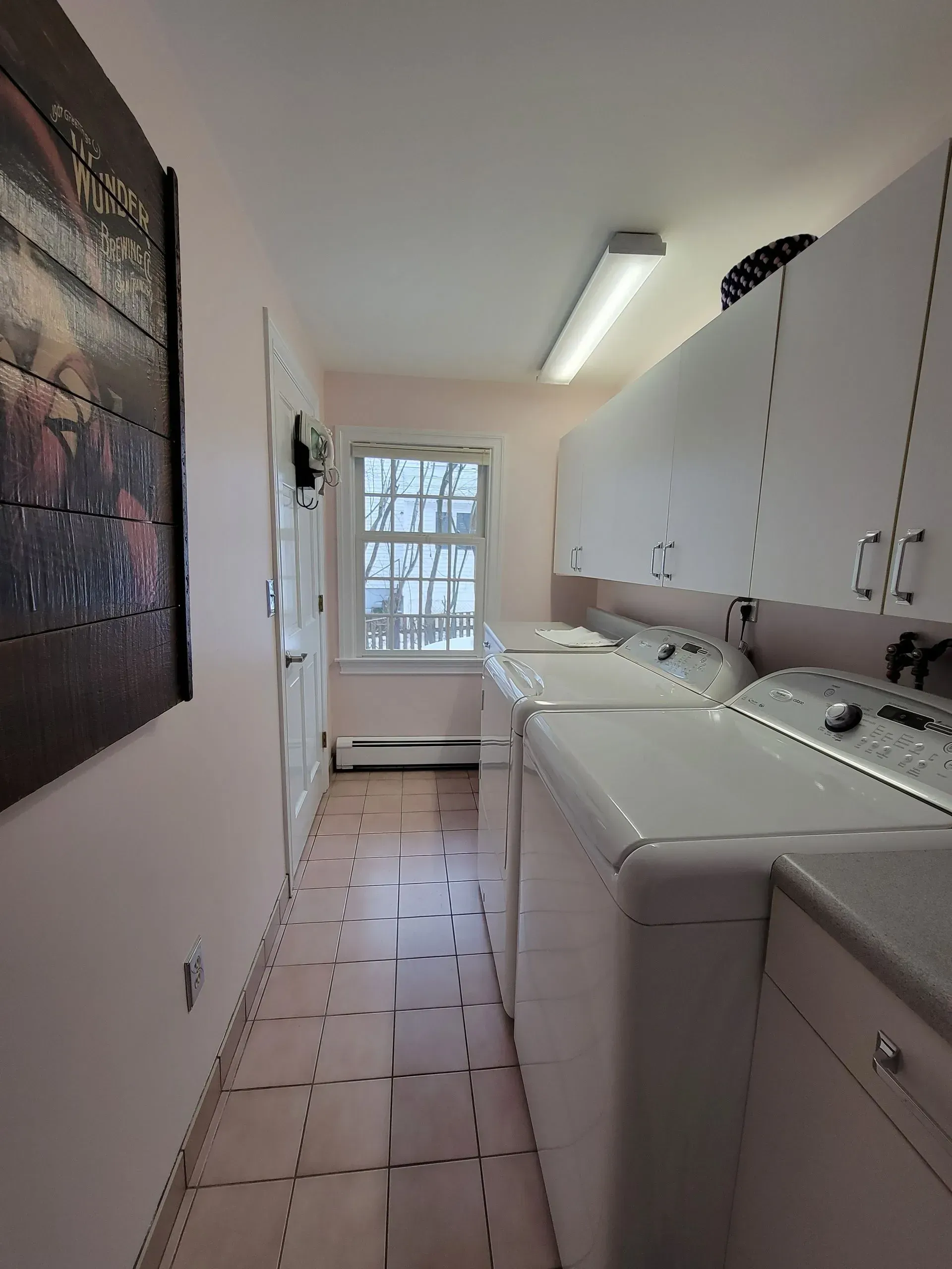 Laundry room with white appliances, pink walls, and upper cabinets. A small window is in the center.