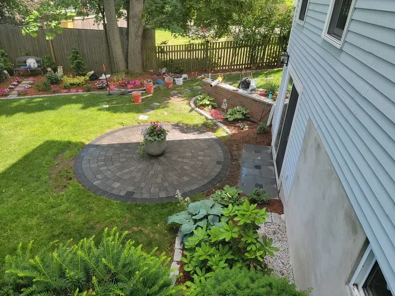 Backyard with a circular patio, lawn, and flower beds, next to a blue house.