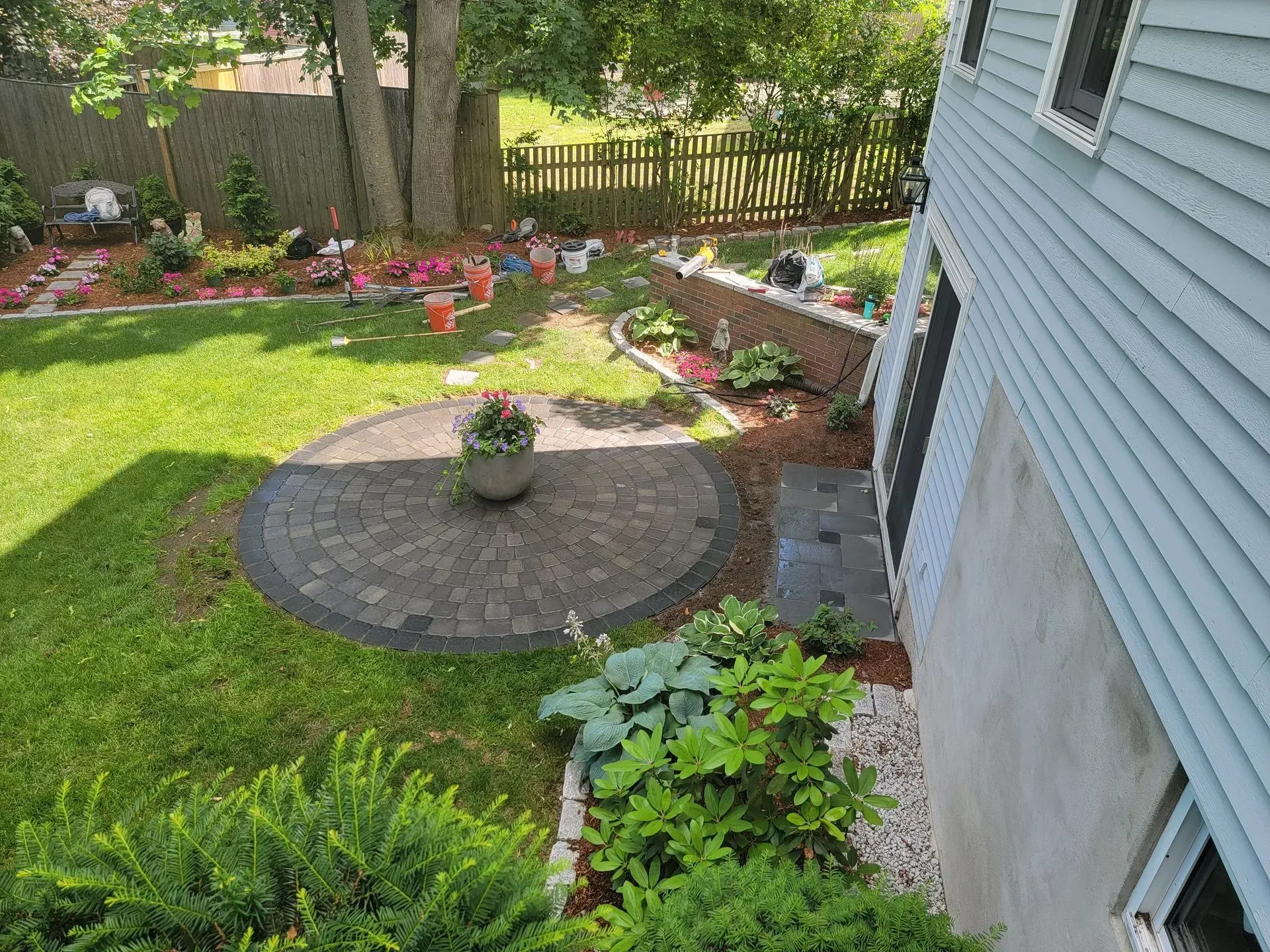 Backyard with a circular patio, flower beds, and a blue house. Green grass and trees are also present.