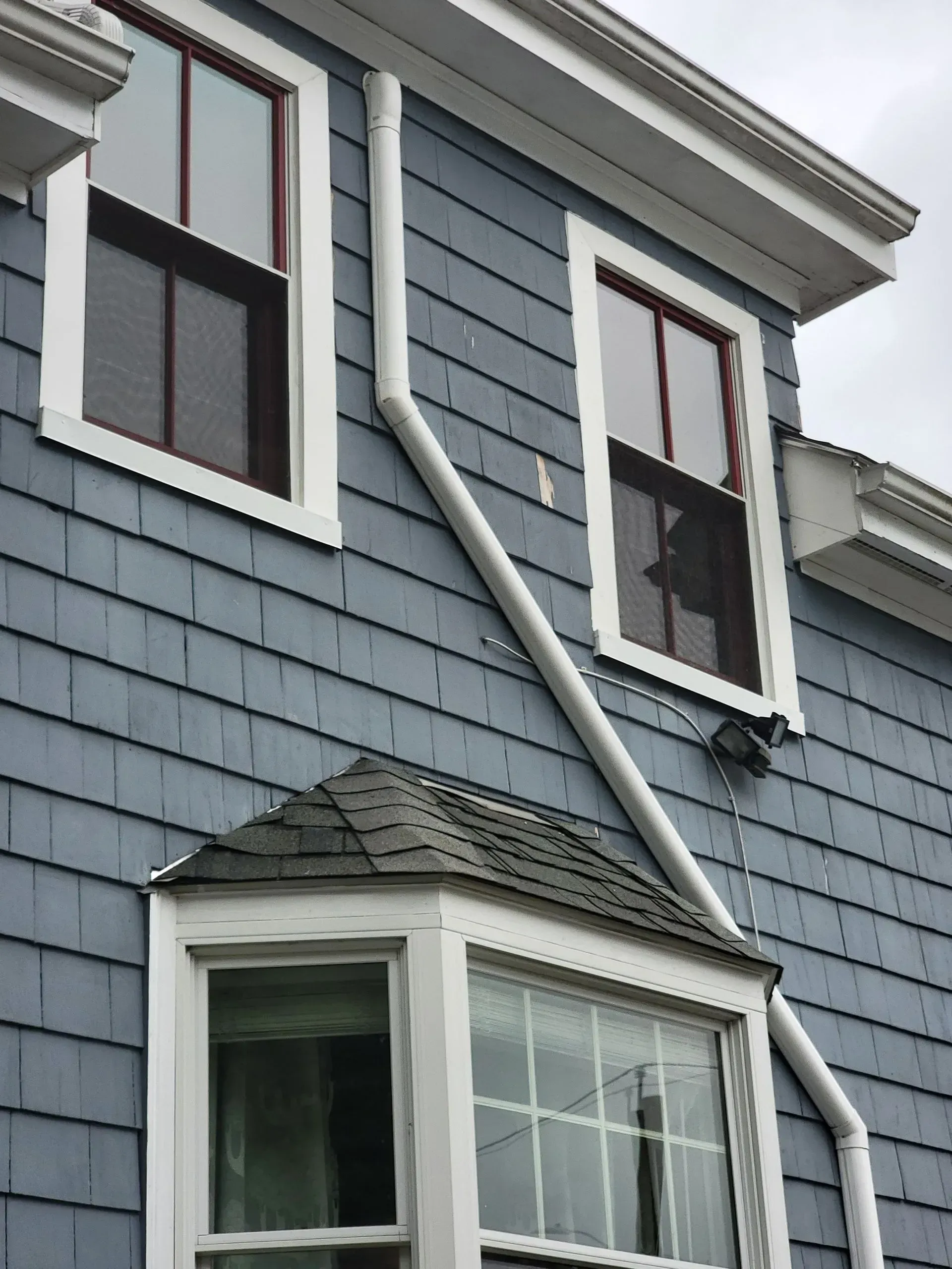 Blue-shingled house with white-framed windows, a bay window, and white downspouts against a gray sky.