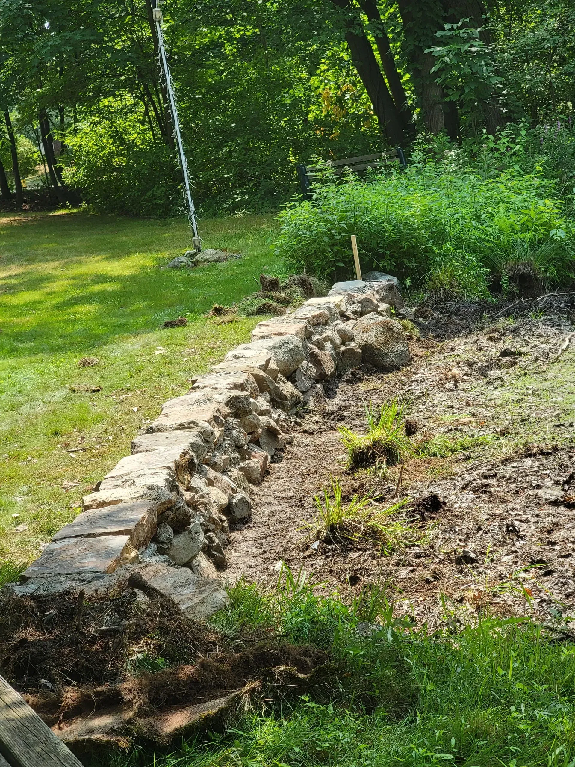 Stone retaining wall in a yard, soil and grass present, woods in background.