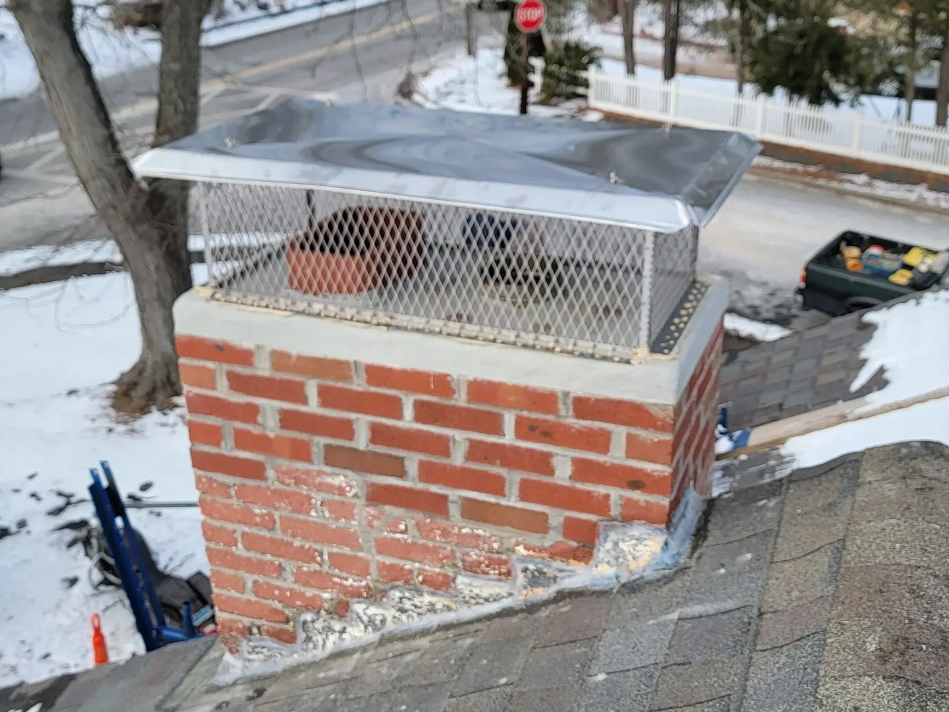 Brick chimney with a metal cap on a rooftop, with snow and a road in the background.