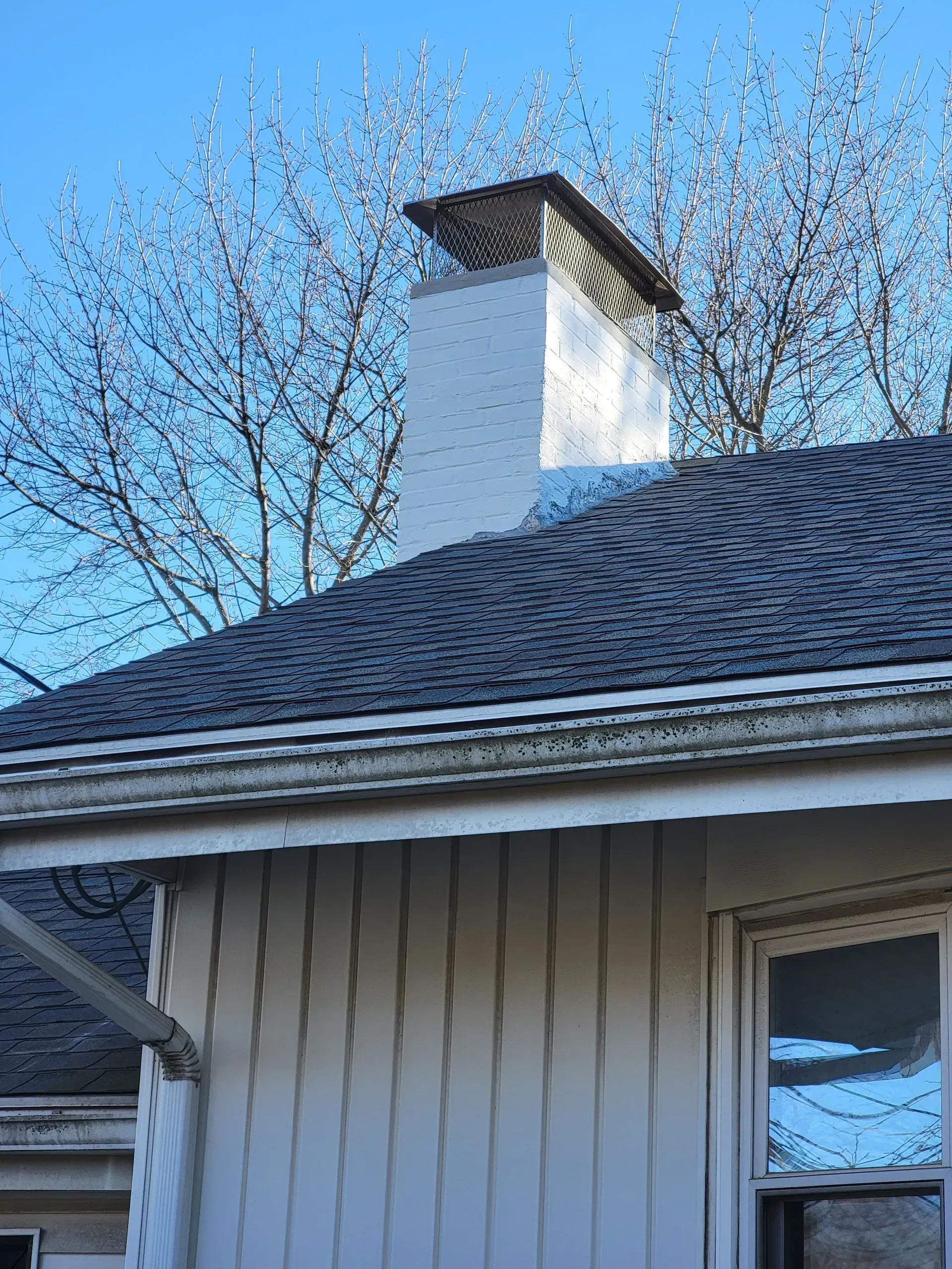 White chimney on a dark roof with a metal cap, trees in background, and a white house with a window.