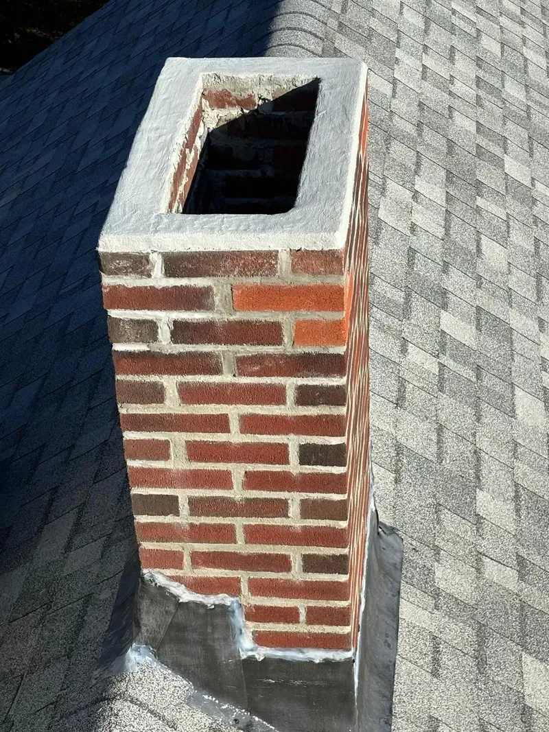 Brick chimney on a gray shingle roof, with a rectangular opening and a concrete cap.