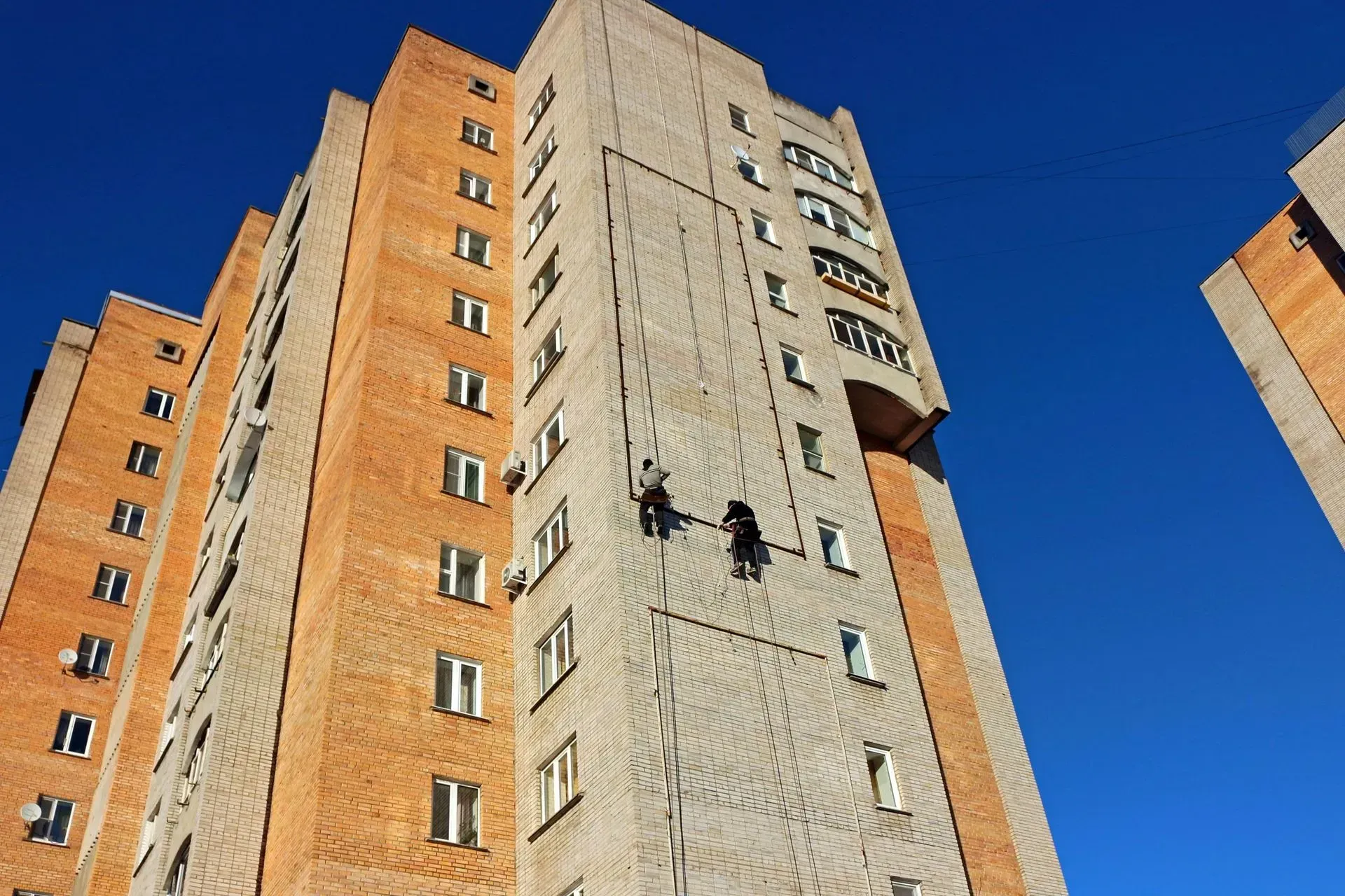 Two window washers on ropes, cleaning a tall brick and concrete apartment building under a blue sky.