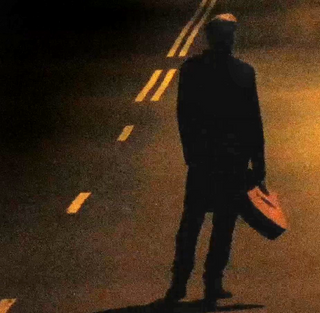 A still from professional videography by Stu Spence depicts a lone man carrying a guitar down a dimly lit road