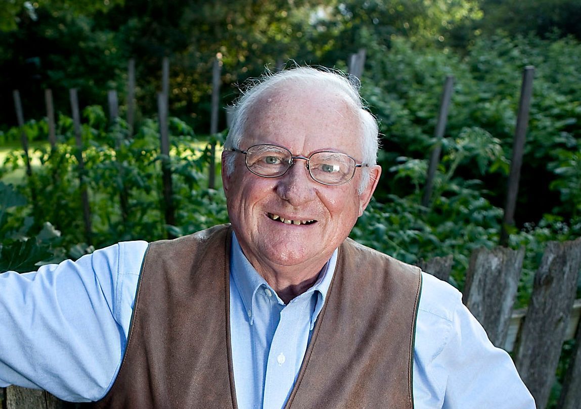 An elderly man wearing glasses and a vest is standing in front of a wooden fence.