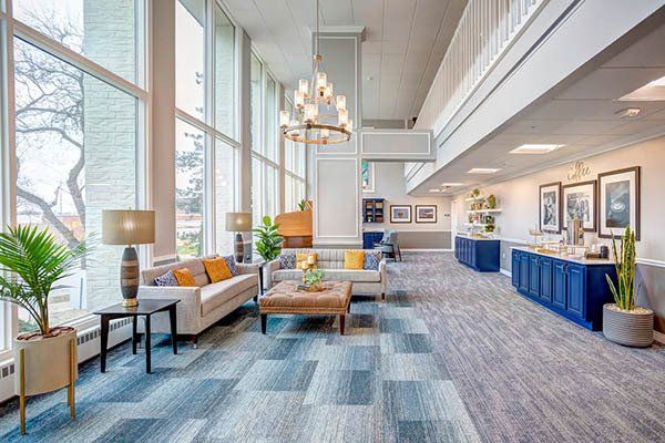 Lobby with large windows, light sofas, and blue cabinets.  Carpeted floor, neutral tones.  Chandelier and plants.