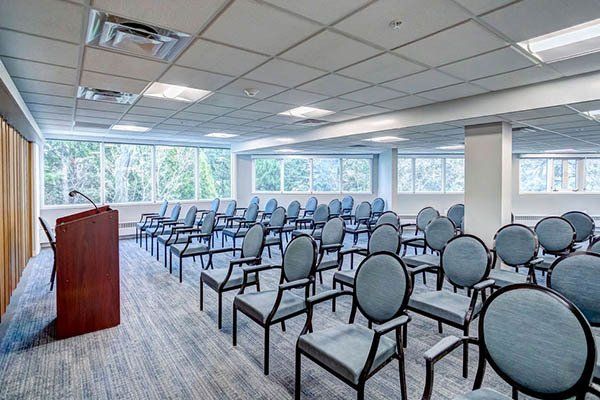 Empty conference room with rows of blue chairs facing a wooden podium; large windows in the background.