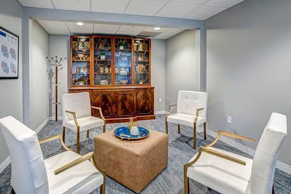 A waiting room with four white chairs around an ottoman, and a wooden cabinet filled with items.