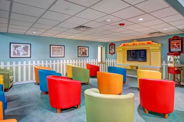 Small colorful theater room with rows of rounded chairs facing a stage. White picket fence decorates one side.