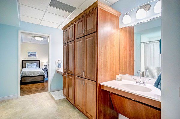 Bathroom with tall wood cabinets, vanity, and a doorway to a bedroom with a bed. Light blue walls.