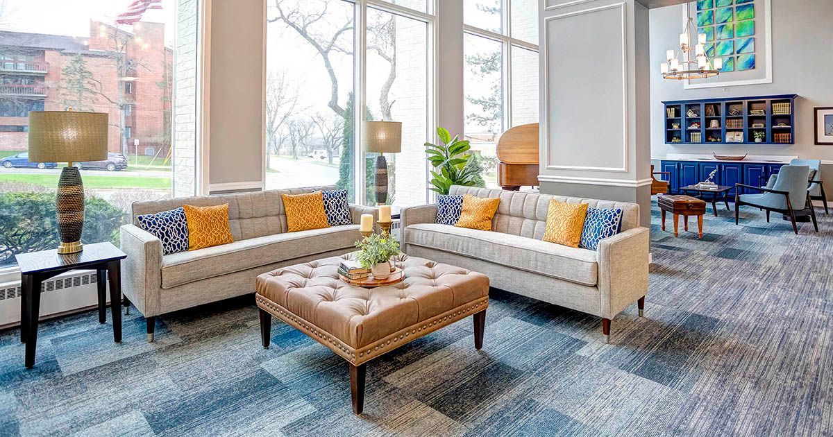 Lounge area with gray sofas, patterned rug, large windows, and a tufted ottoman in a building.