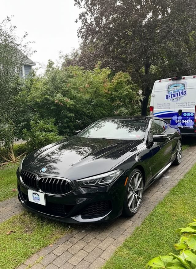 Black BMW coupe parked on a brick driveway beside a van with a company logo.