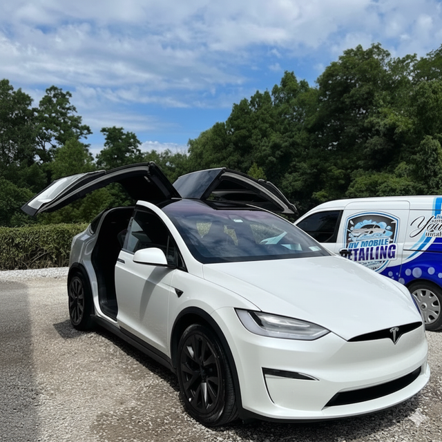 White Tesla SUV with falcon wing doors open, parked near a detailing van. Blue sky and trees in the background.