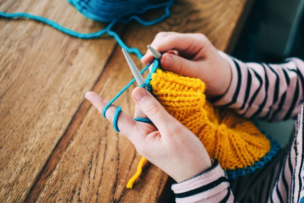 Hands knitting with blue and yellow yarn on a wooden table.