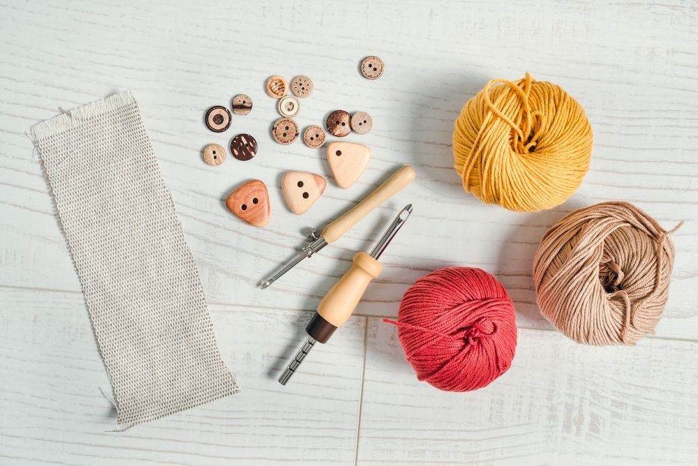 Crafting supplies on a white wooden surface: yarn balls, buttons, punch needles, and fabric.