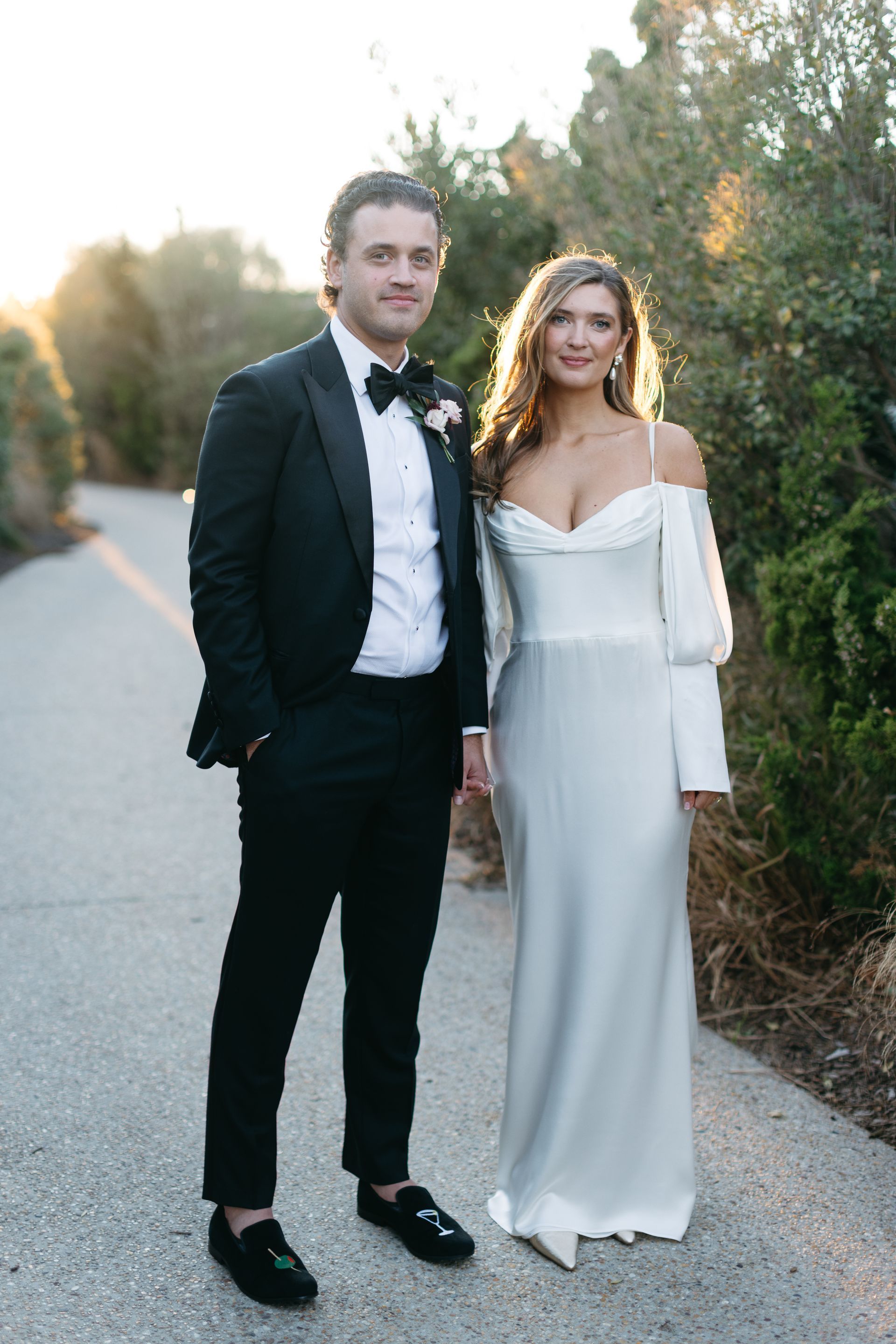 Groom in black tuxedo and bride in white gown pose on a pathway; golden hour lighting.
