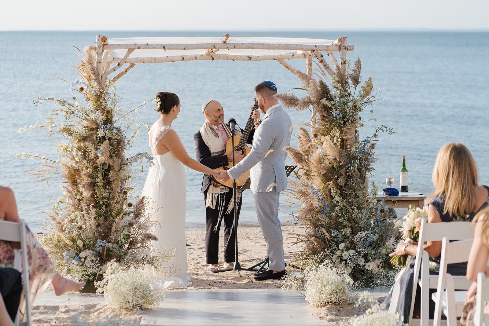 Wedding ceremony on beach: Bride, groom holding hands under decorated arch, officiant in middle.
