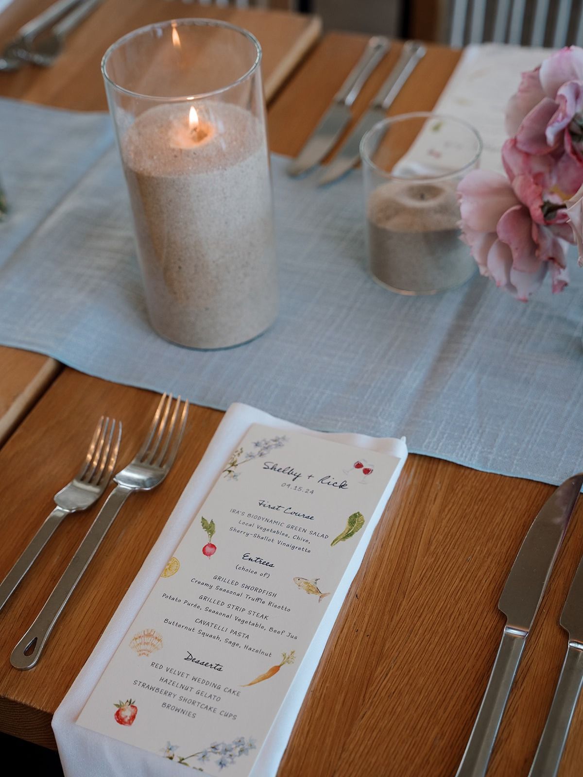 Table setting with menu, candle, flowers. Light blue runner, wooden table.