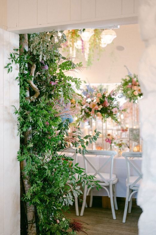Greenery frames doorway to a wedding reception: floral arrangements, white chairs, and table settings.