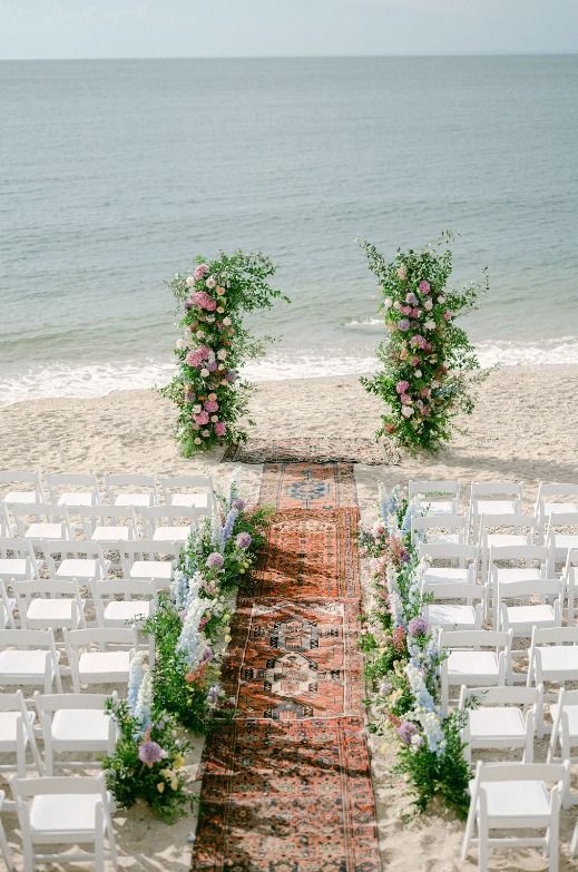 Wedding ceremony on a beach with an archway of flowers, rug aisle, and white chairs facing the ocean.