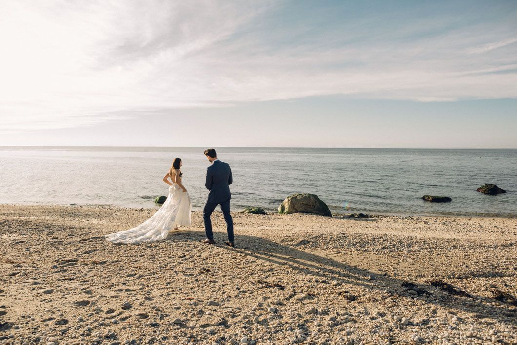 Bride and groom stand on a beach. The bride is in a white dress, and the groom in a suit, the ocean in the background.