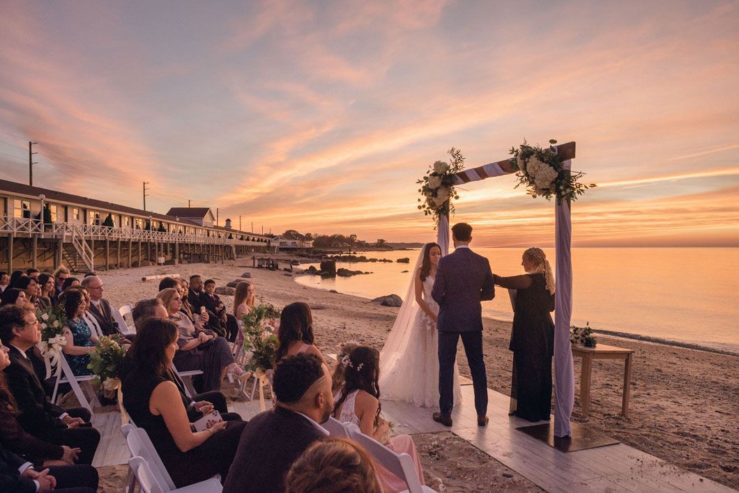 Couple at beach wedding ceremony under an arch, sunset backdrop. Guests seated.