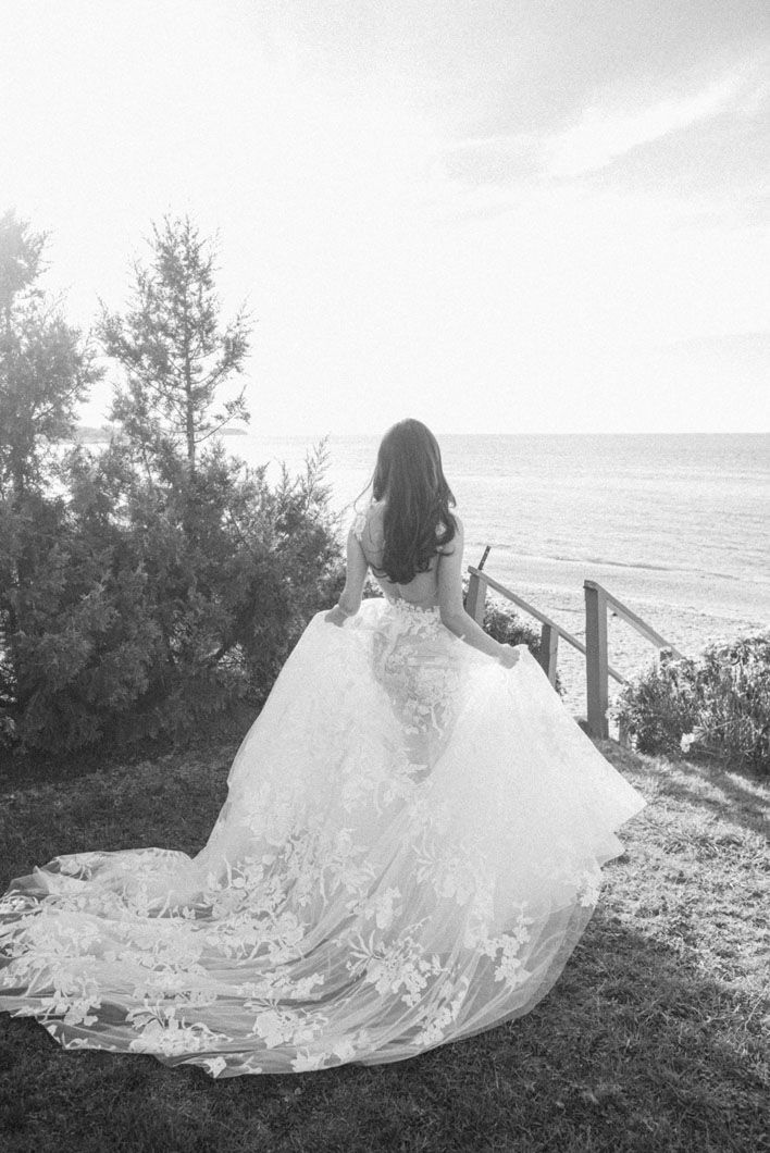 Bride in wedding dress, back to camera, overlooking ocean, train flowing, sunny day.