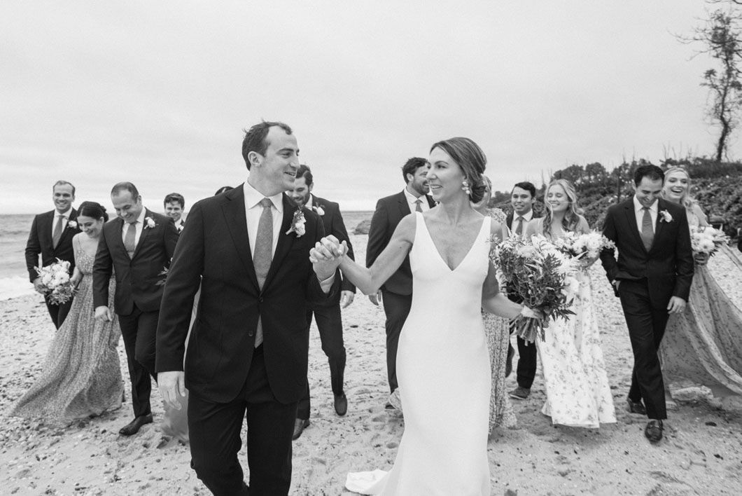 Bride and groom walk on beach, holding hands, followed by wedding party. Black and white.