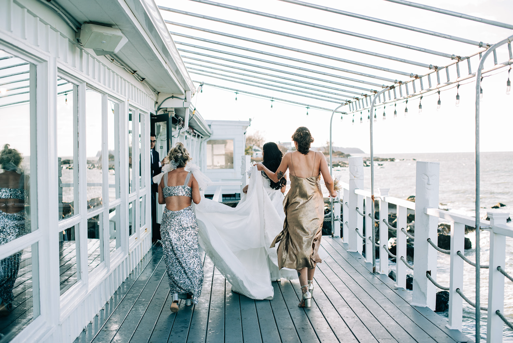 Bride with two bridesmaids walking on a wooden deck, ocean view, sunlight.