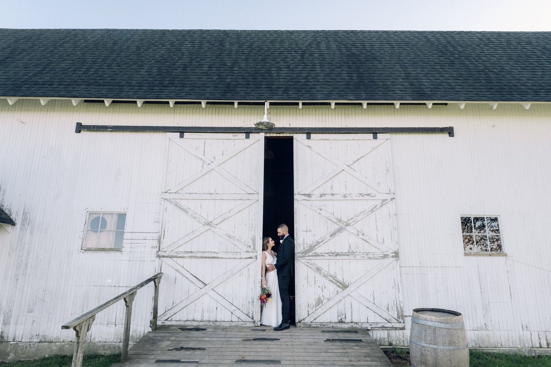 Bride and groom stand framed in the doorway of a white barn, looking at each other.