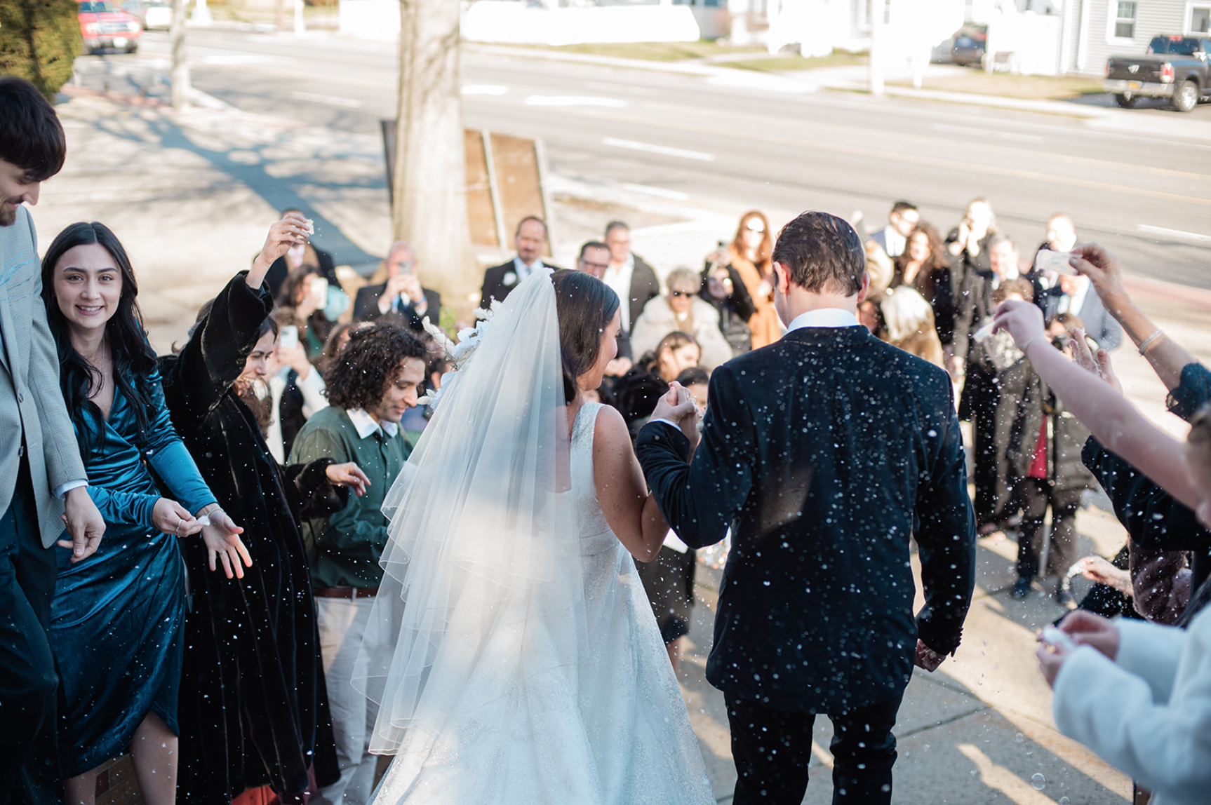 Newlyweds walk amid confetti thrown by guests outside a building; sunny day.