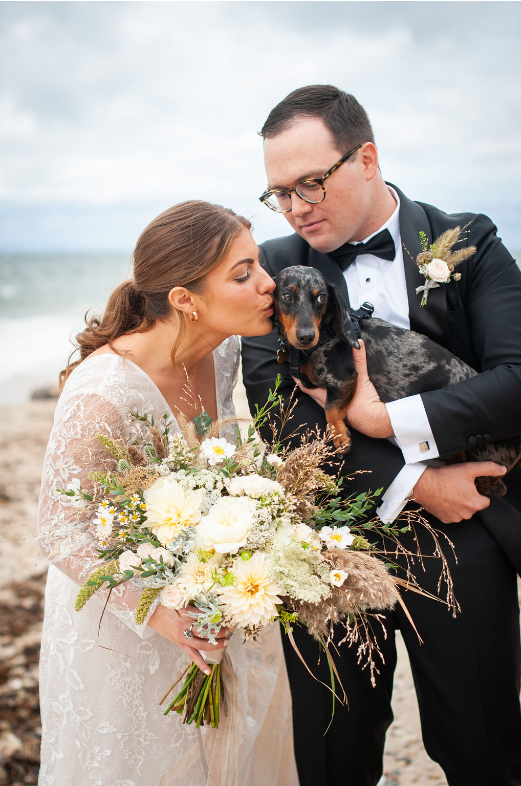 A bride and groom are kissing their dog on the beach.