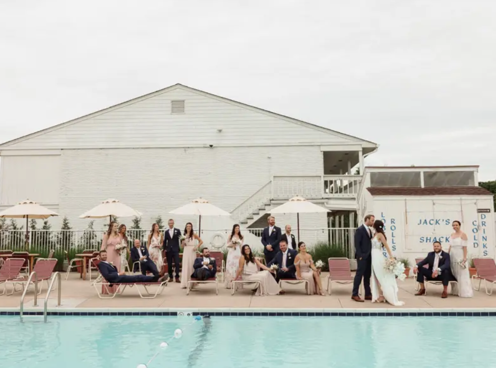Wedding party poses by a pool; people in formal attire, white building backdrop.