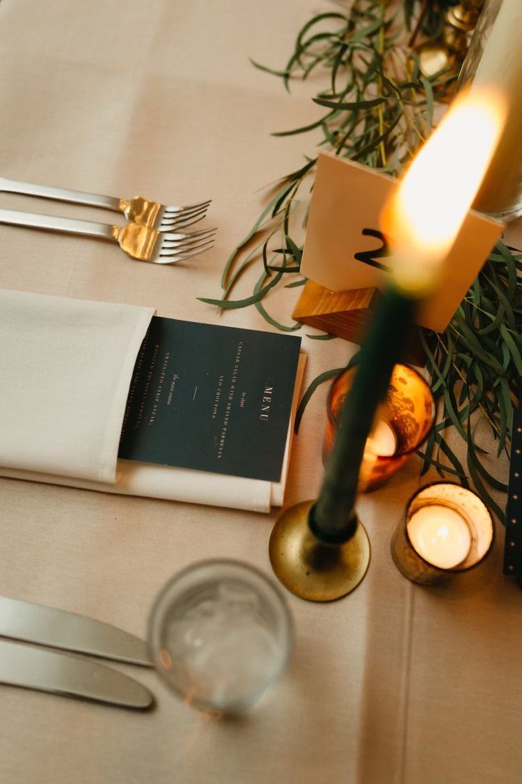 Table setting with lit candle, napkin, utensils, place card, greenery.