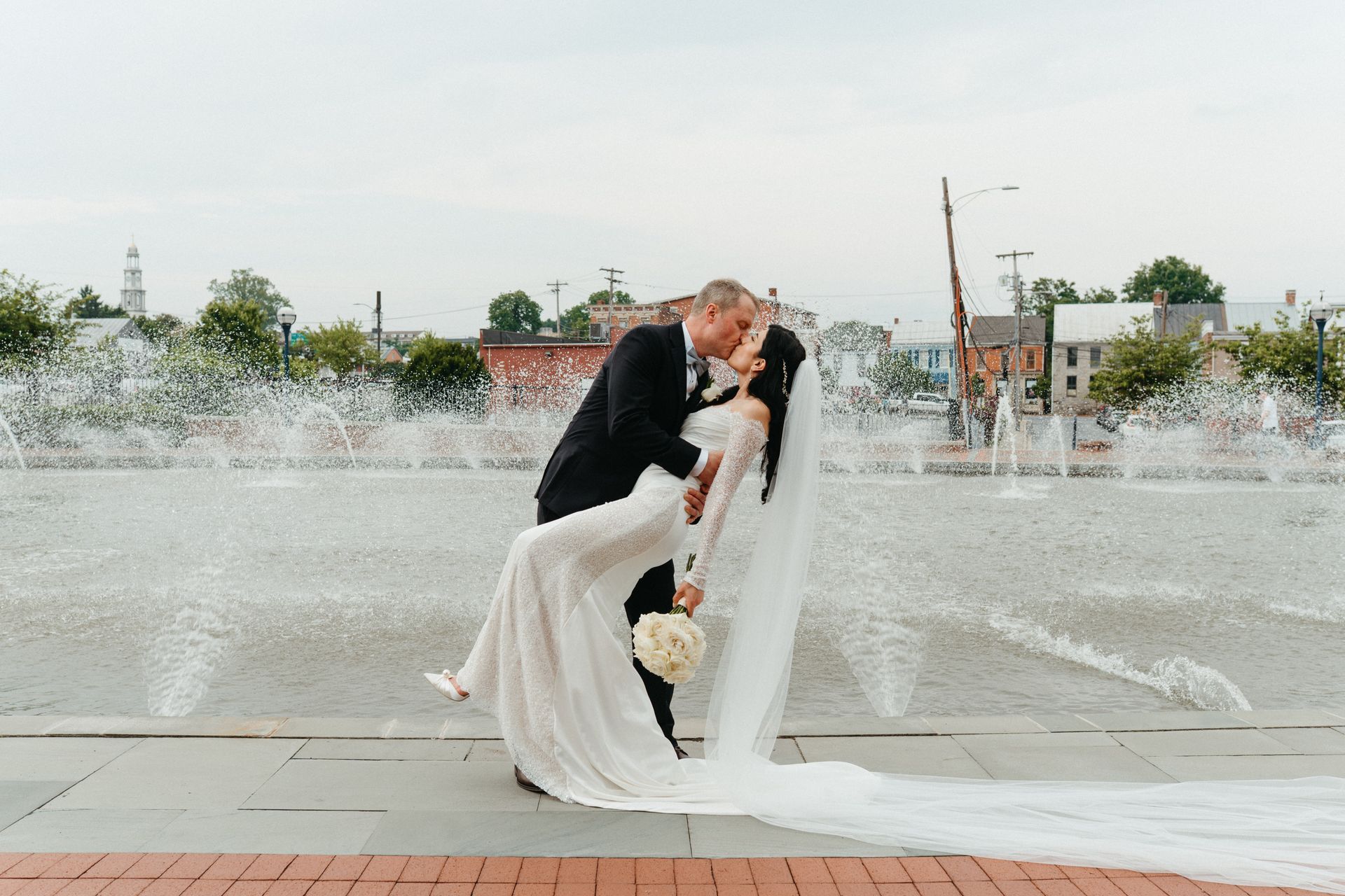 Groom dips bride for a kiss in front of a fountain. She wears a veil and holds a bouquet.