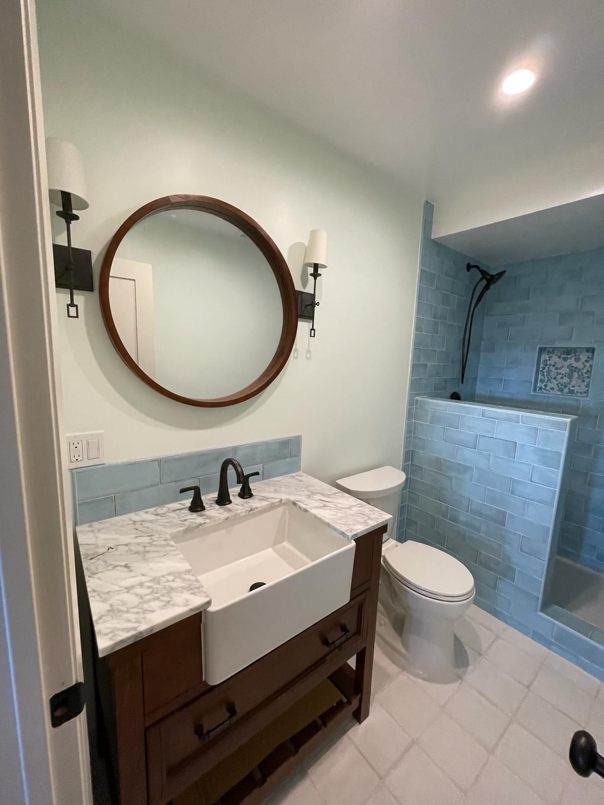 Bathroom with a brown vanity, white sink, round mirror, and a blue-tiled shower.