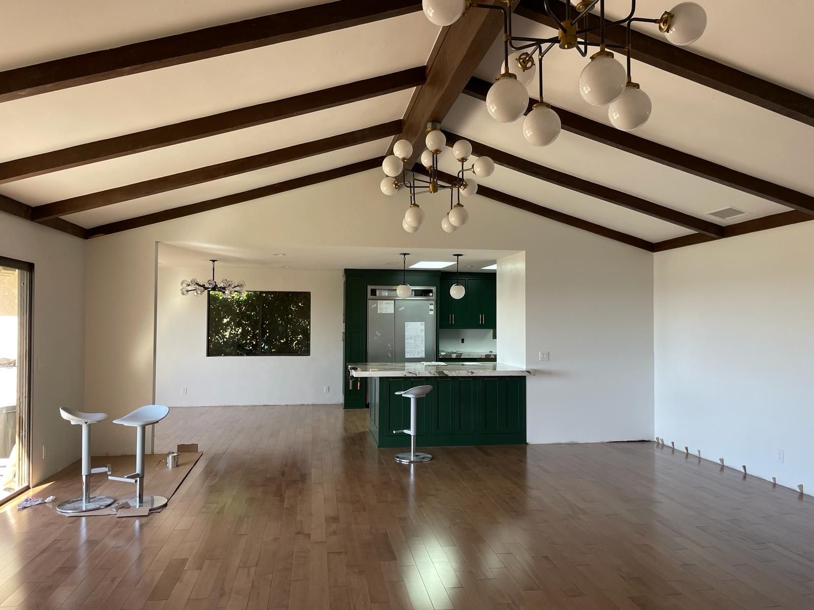 Empty living room with green kitchen, wood floors, exposed beams, and modern chandeliers.