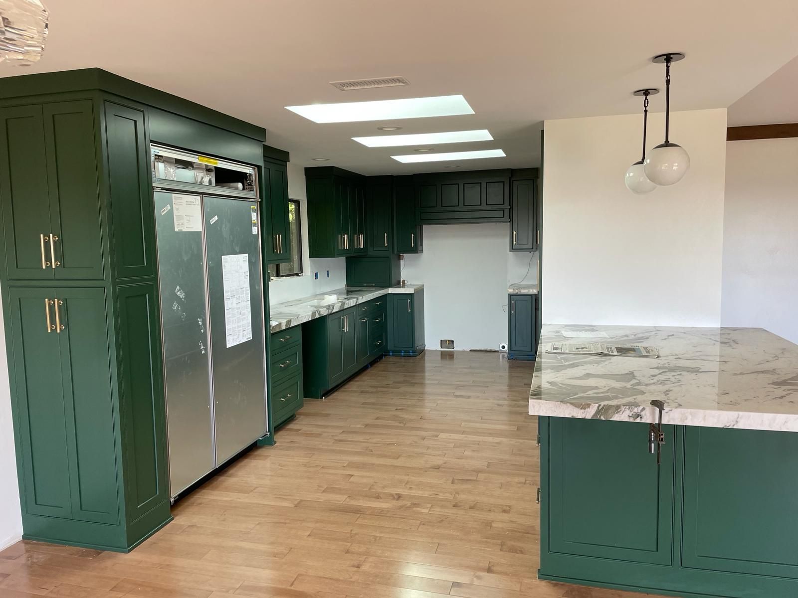 Green-painted kitchen cabinets and island with white countertops. Stainless steel refrigerator and globe pendant lights.