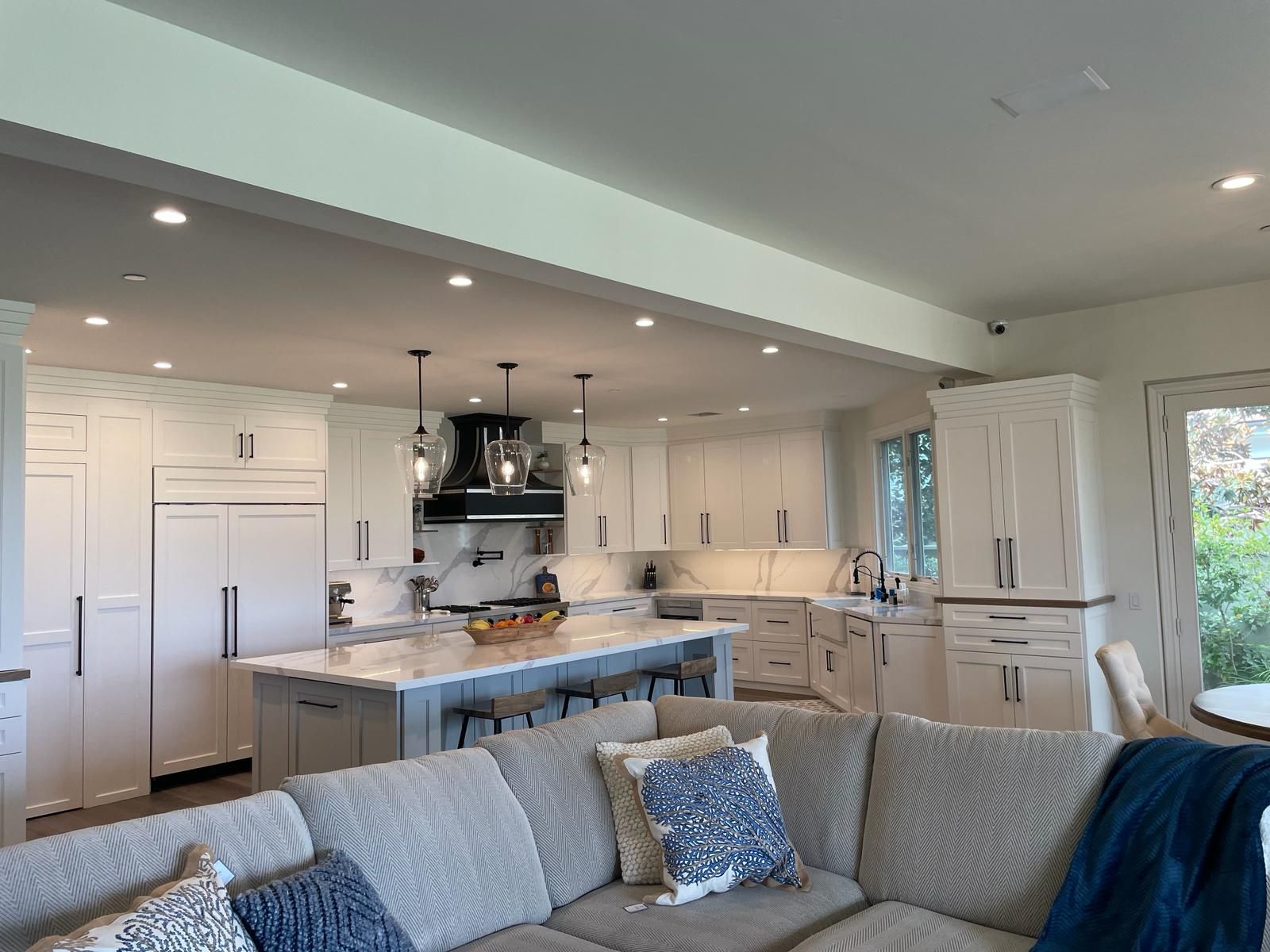 Spacious white kitchen with island, seating, and overhead lighting, visible from living room with gray sectional sofa.