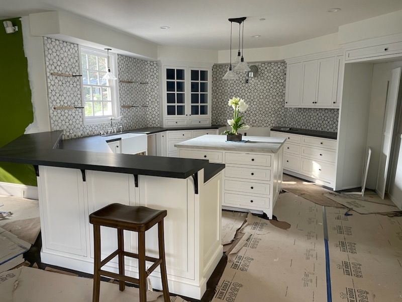 A kitchen under construction with white cabinets, dark countertops, patterned tile backsplash, and a central island.