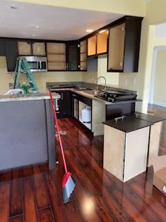 Kitchen under renovation: dark cabinets, exposed interiors, countertop, new flooring, broom, ladder.