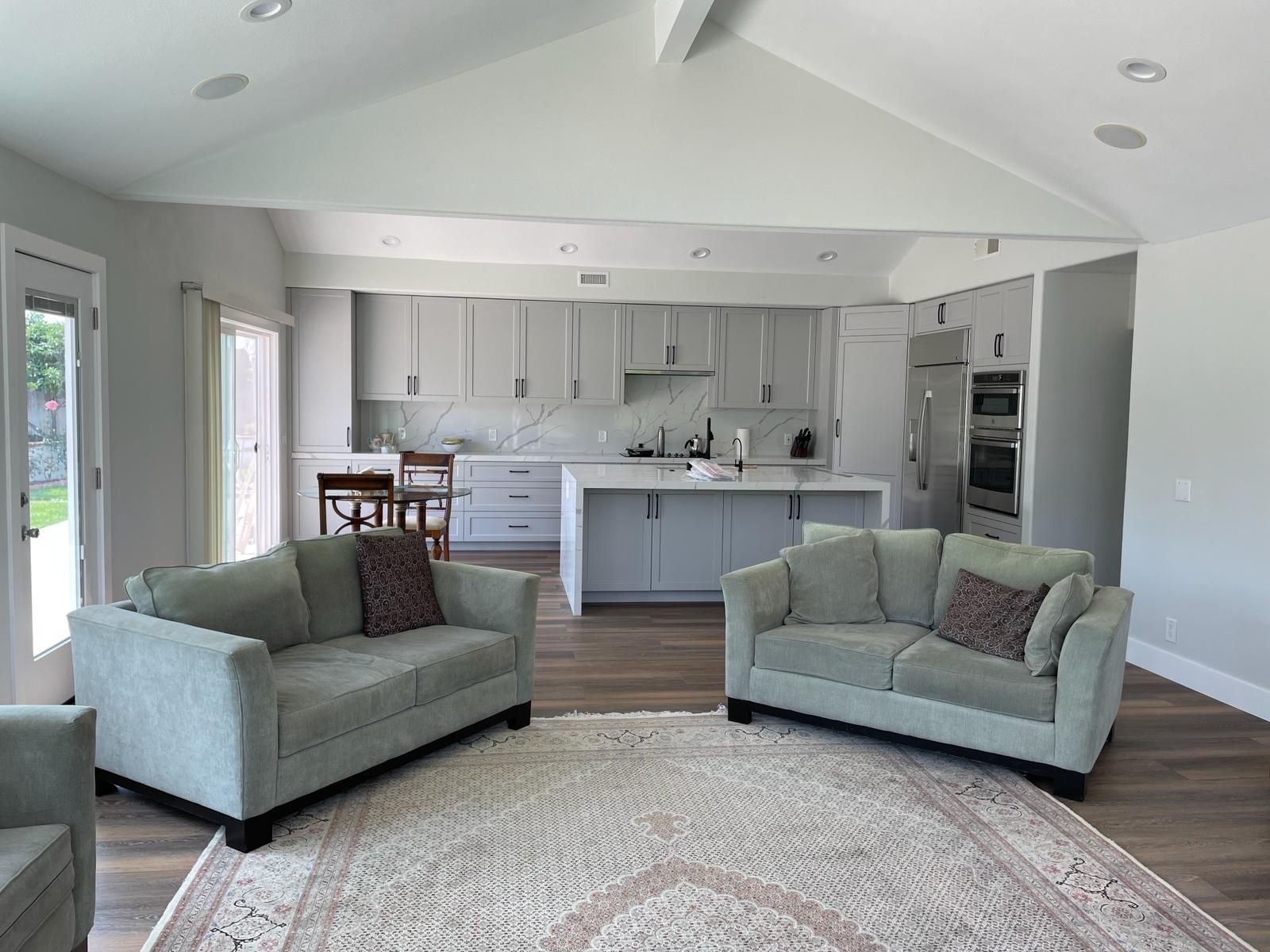 Open-concept living space with two green sofas, a kitchen with gray cabinets, and a rug.