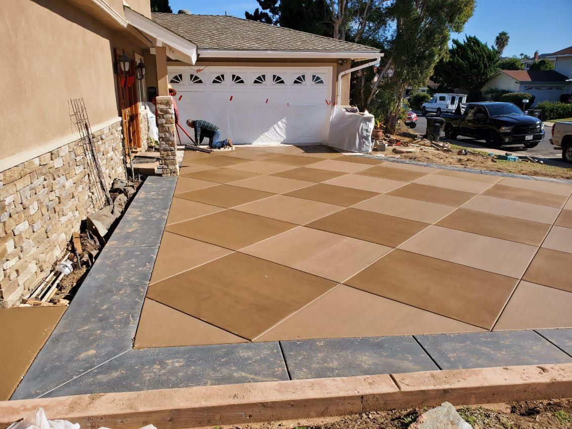Driveway paved with diamond-patterned tan and brown tiles, bordered by dark gray blocks. Person near garage.
