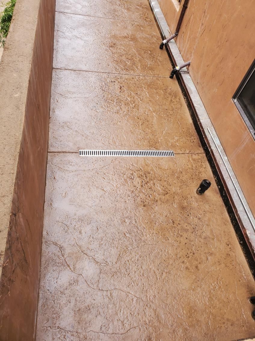 Long, narrow outdoor space with a patterned concrete floor, and walls on either side, with a drainage grate.