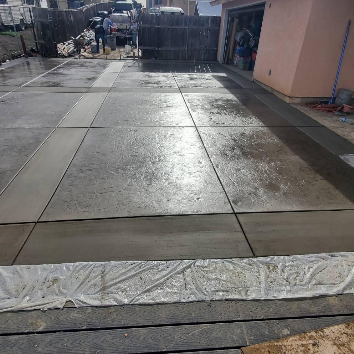 Newly poured concrete driveway with fresh water droplets. A person and construction materials are visible in the background.