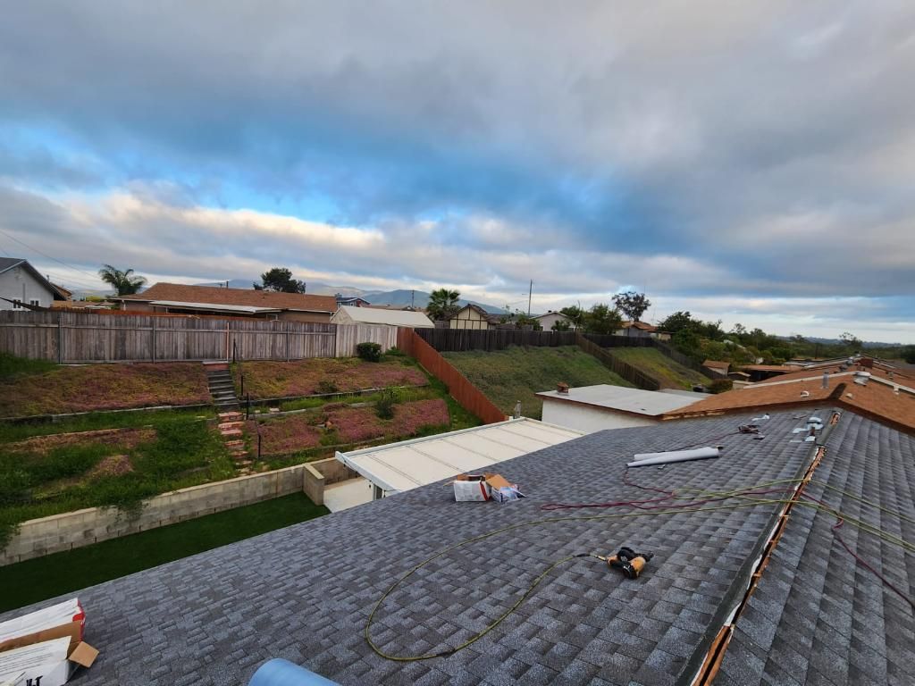 Rooftop view of houses, yards, and sky with clouds. Grey asphalt shingles and brown fence are visible.