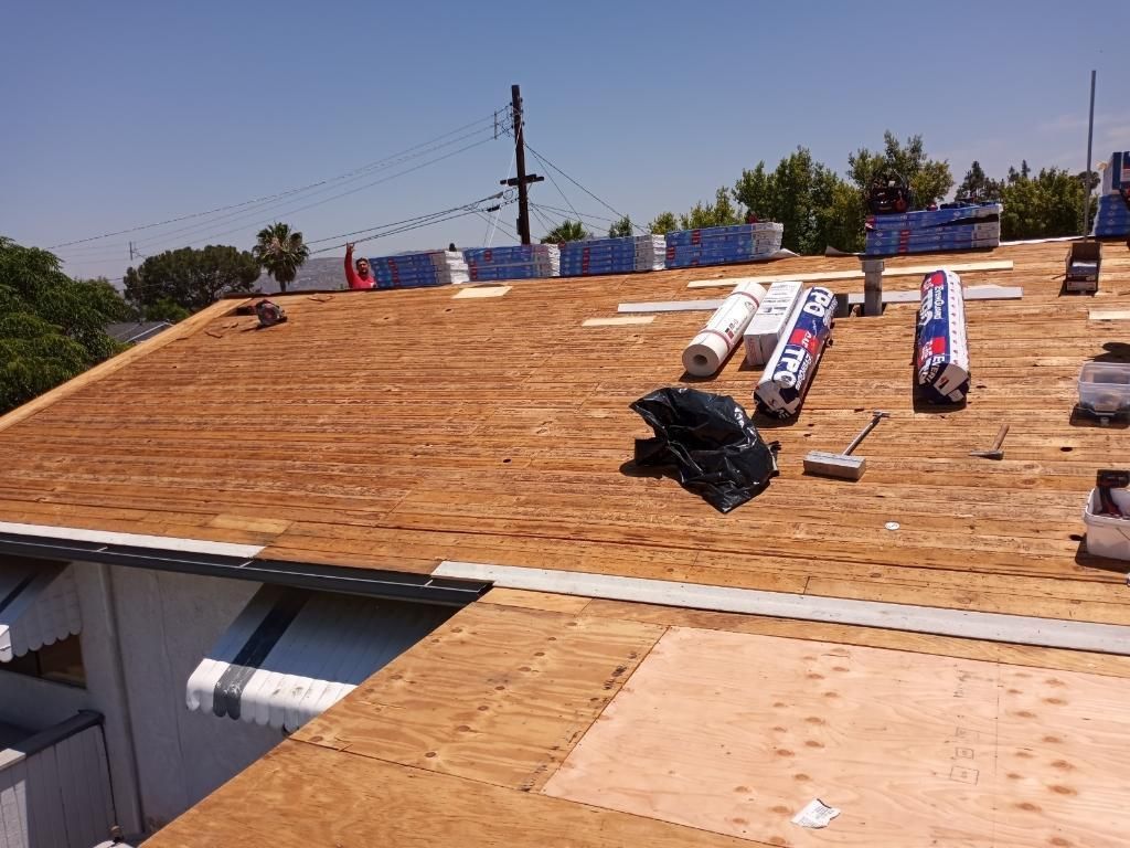 Roof being replaced; plywood, shingles, materials, and workers on the roof against a sunny sky.