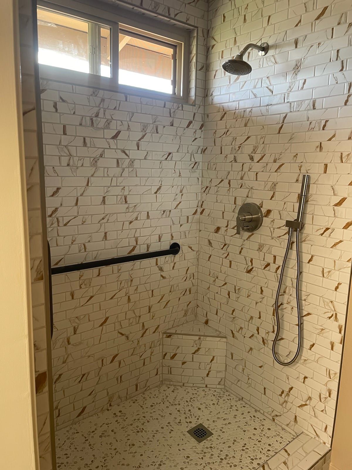 Shower stall with white and gold patterned tiles, grab bar, showerhead, and a small built-in seat.